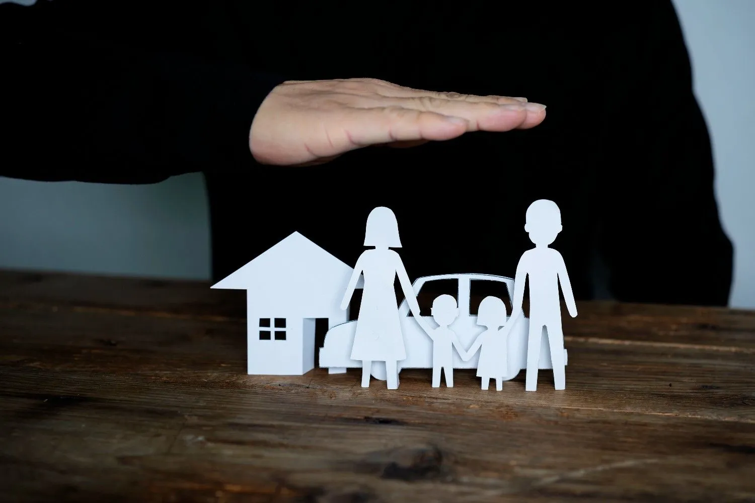 A hand held protectively over paper cutouts of a family, a house and a car
